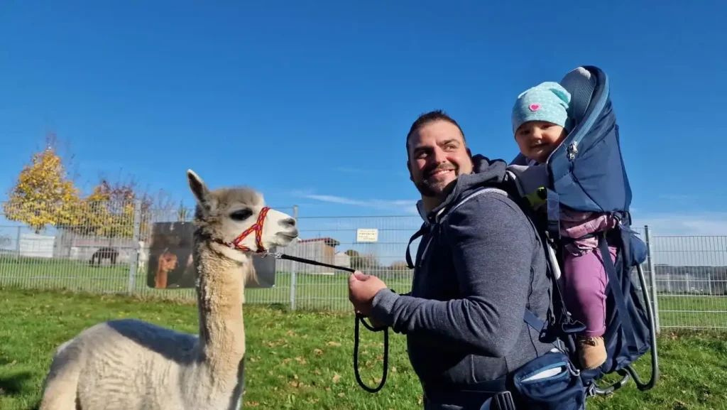 Zu sehen ist ein Mann der glücklich zu seinem Kind auf der Rückentrage schaut. Dabei hat er unser Alpaka "Luca" bei der Hand